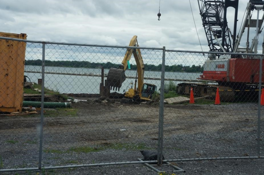 View of a construction site at the waterfront through a fence. An excavator is down near the water level and using a bucket to remove concrete from the boat ramp. There is also a crane sitting in the parking lot nearby.