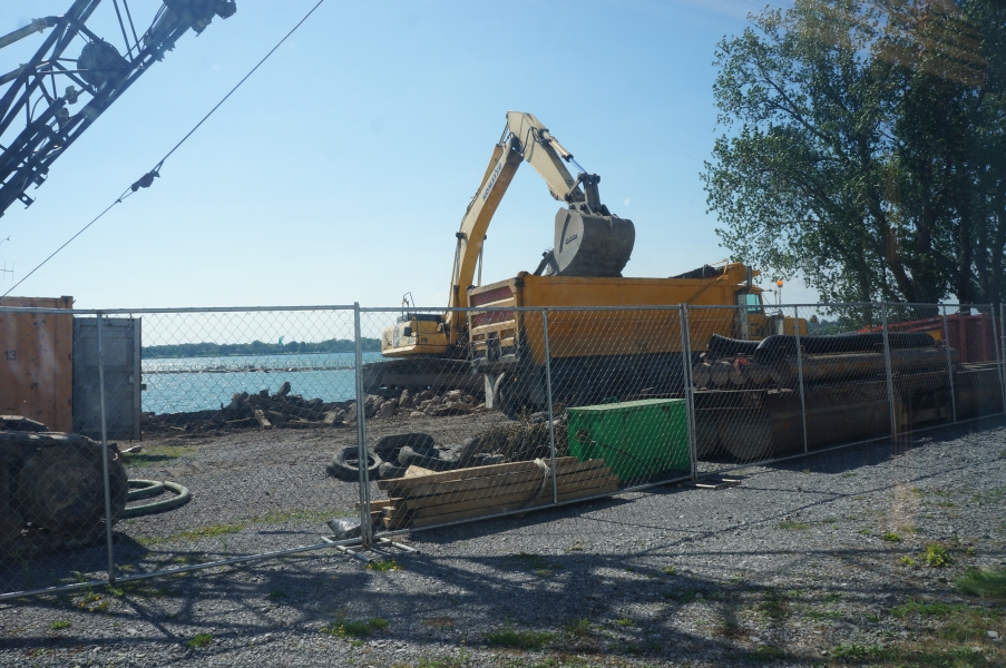 View of a construction site through a fence. An excavator is dumping debris into a dump truck.