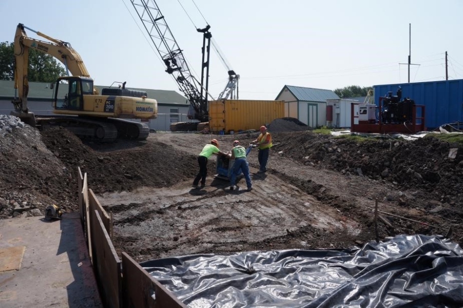 Three construction workers on a slope move a small heavy machine that is pounding dirt flat. An excavator sits at the top of the slope.