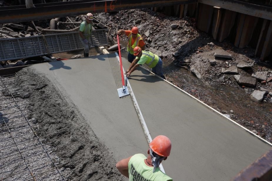 Four workers at a construction site use a tool to put lines on fresh smooth cement.