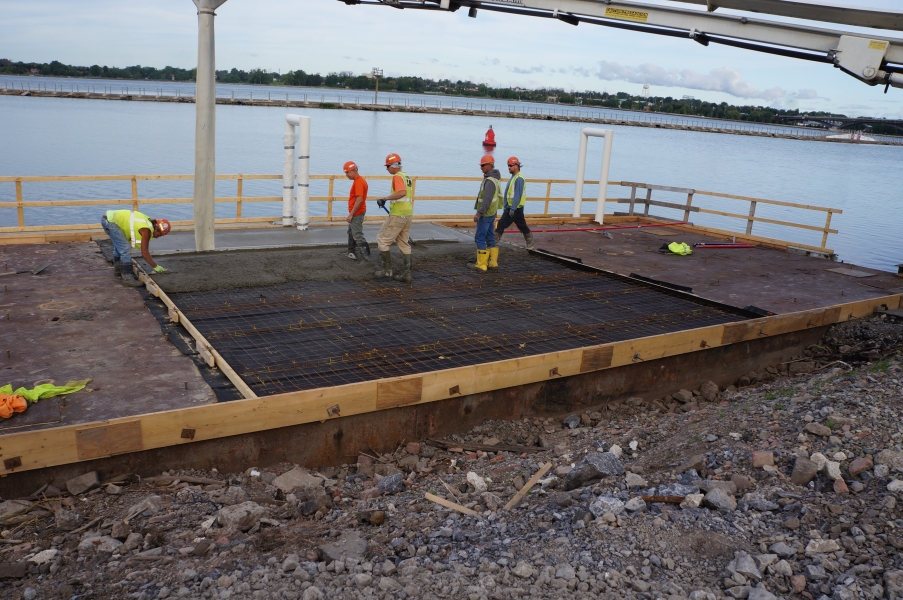 Construction workers spreading cement on a frame of rebar on top of a metal surface by the water.