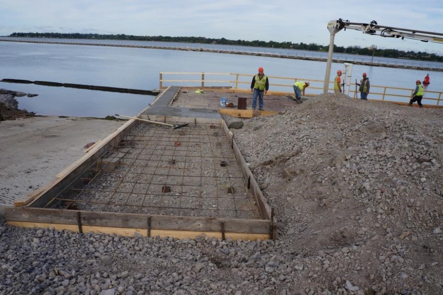 A construction site with workers by the waterfront. A frame with rebar and gravel goes down to a dock next to a boat ramp.