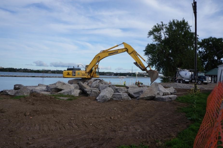 An excavator moves larges stones near the waterfront. A pile of large stones sits on the ground in front of the excavator.