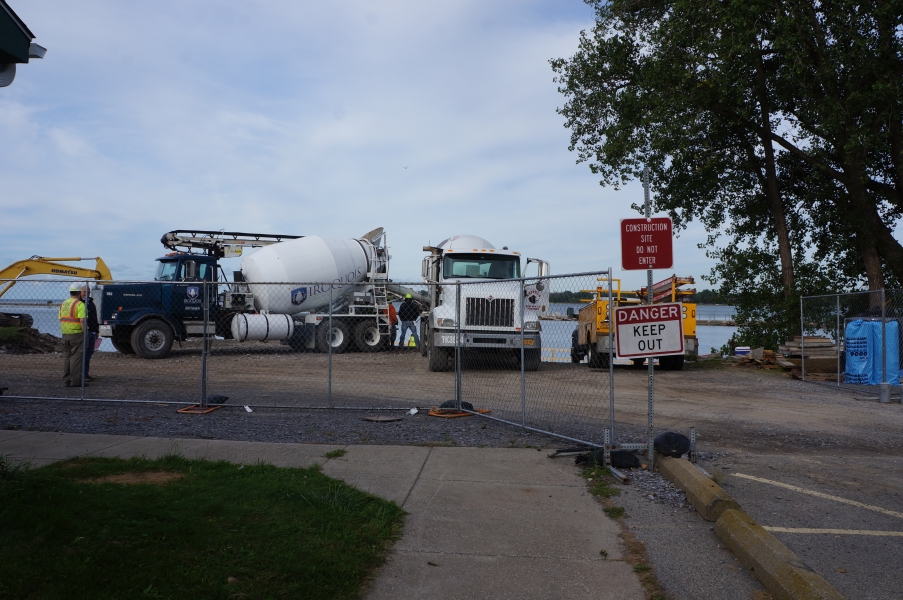 A fence with a sign "DANGER KEEP OUT" blocks a construction zone with two cement trucks and some other equipment. There is a body of water behind the construction zone.