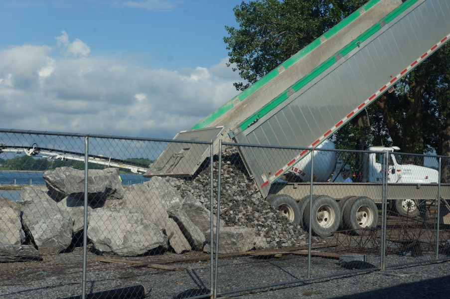 A dump truck pours out a load of small stone next to a pile of boulders.