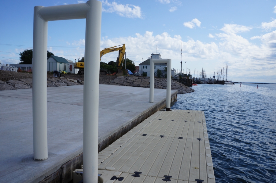 A concrete dock with two white metal u-shaped bars. A plastic platform floats in the water next to the dock, attached to the metal bars. An excavator works in the distance.