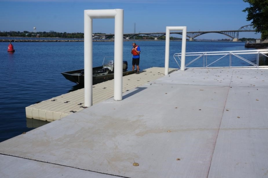 A person stands by a boat on a floating dock by the water. The main part of the dock is concrete and two white metal u-shaped bars connect to the floating dock.