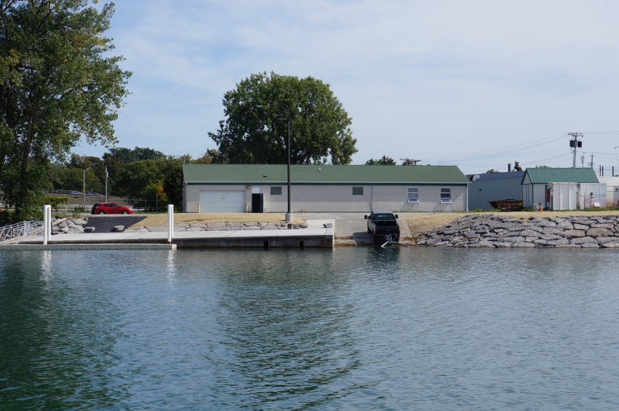 View from the water of a one-story building with a dock and ramp. The shoreline has neat stonework and the dock has a floating dock. A truck and trailer are backed down the boat ramp.