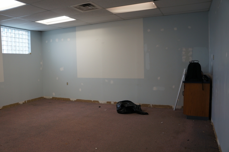 A room being renovated. There are putty patches on the wall and the base board has been removed. There is a bag of trash in the middle of the room.