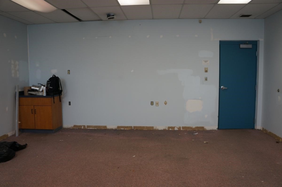 A room being renovated. There are putty patches on the wall and the base board has been removed. There is a small cabinet with a sink in one corner with a back pack and supplies on it.