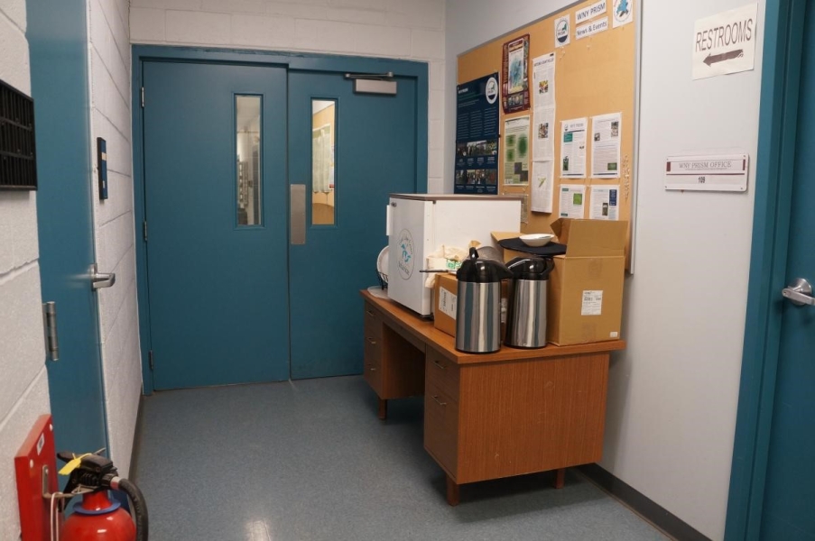 A desk with a mini fridge and other things piled on top of it in a hallway.