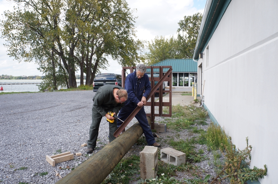 Two people attach a steel structure to a utility pole lying on the ground. One holds the steel while the other uses a drill.