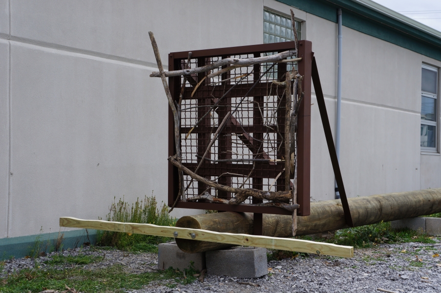 A steel mesh platform at the top of a utility pole lying on the ground. There is driftwood affixed to the frame and a beam across the top of the pole to function as a perch.