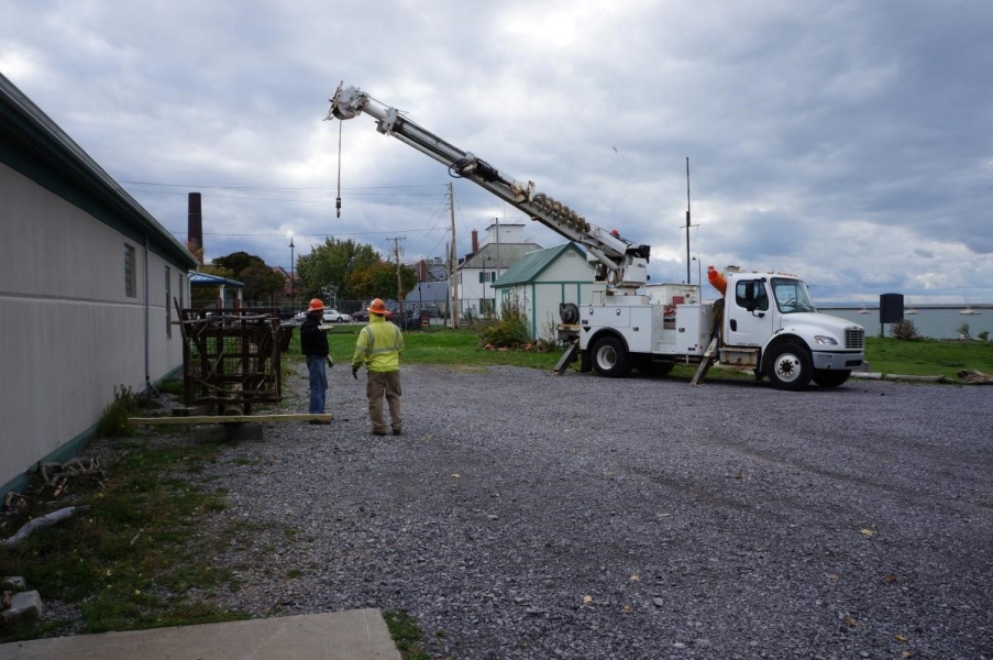 Two people in hard hats stand next to an osprey pole on the ground and a truck with a crane.