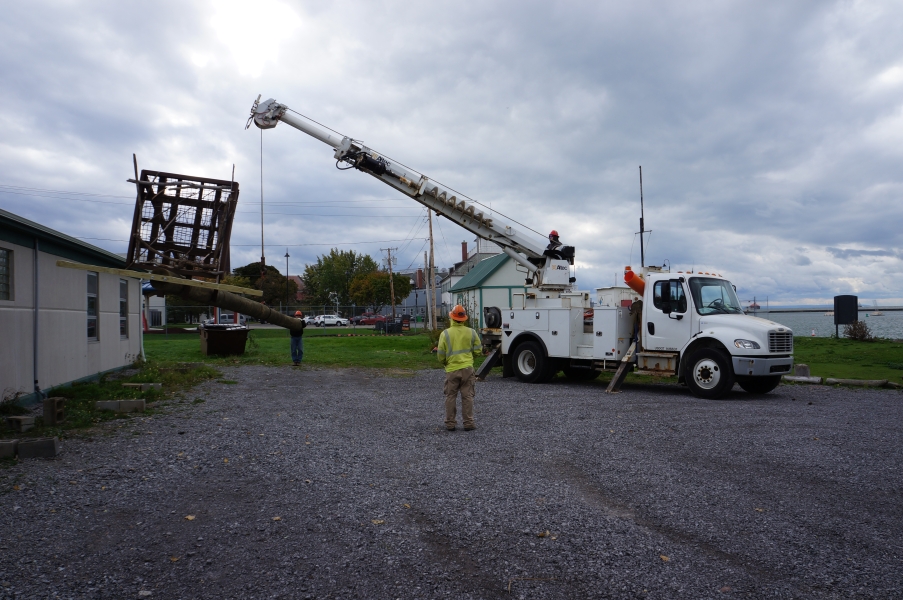 A truck with a crane lifts an osprey pole off the ground while one person steadies one end and another person observes.