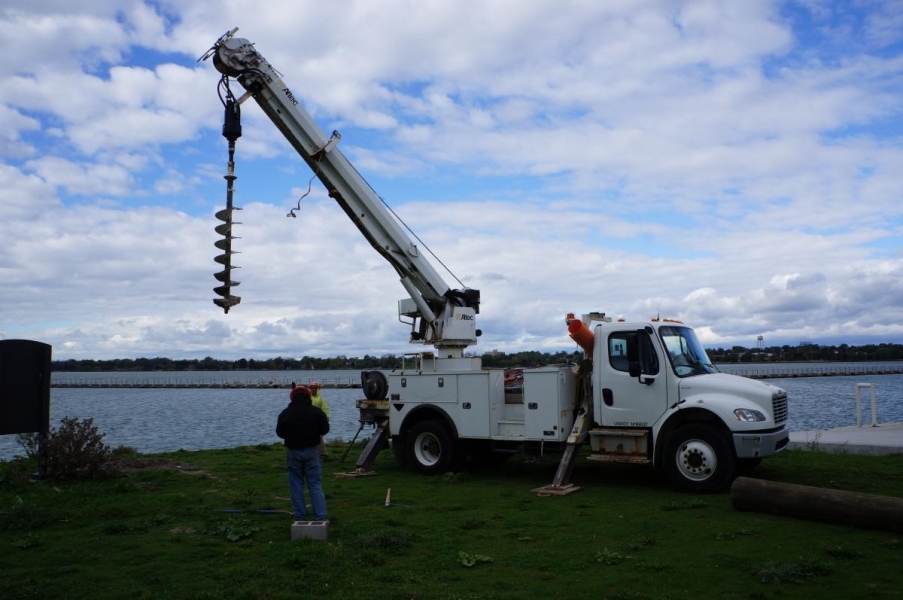 Two workers stand next to a truck with an auger near the waterfront.