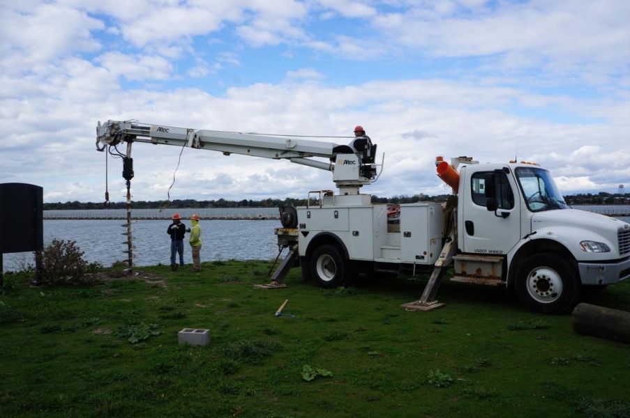 Two workers stand by while a third worker on the truck operates an auger on a truck to drill a hole by the waterfront.