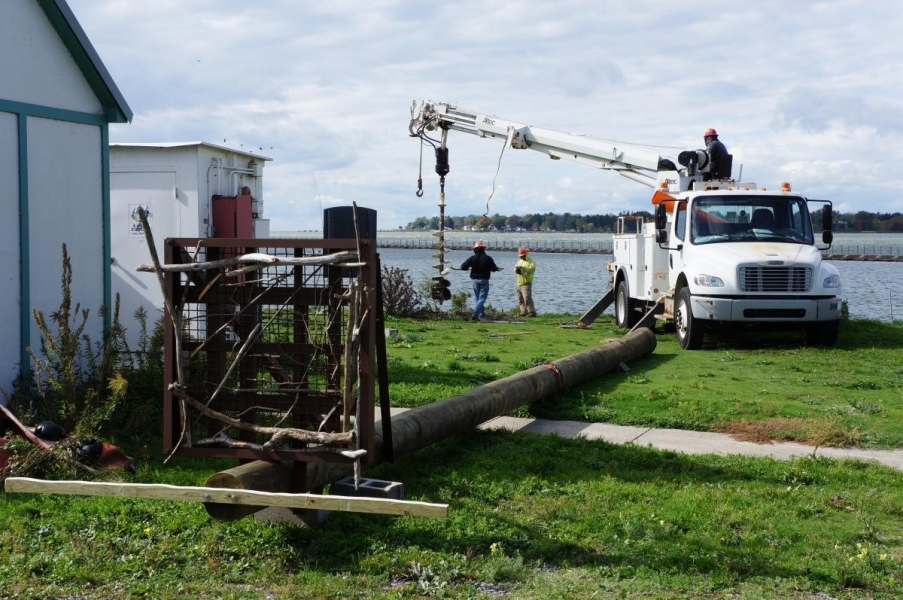 A utility pole with a steel platform lies on the ground near a truck drilling a hole in the ground near the waterfront.