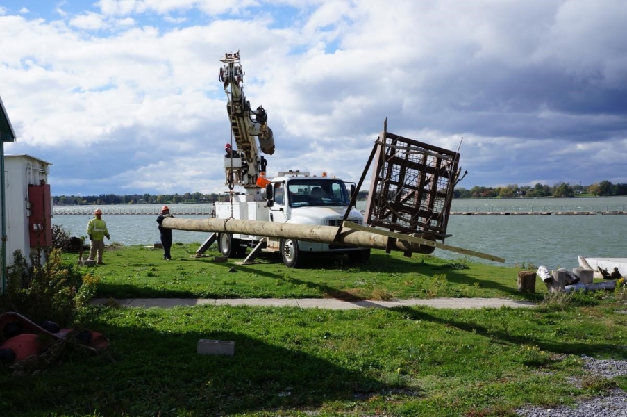 Workers use a truck with a crane to move the osprey pole near the waterfront. The pole is perpendicular to the ground and only a few feet above it.