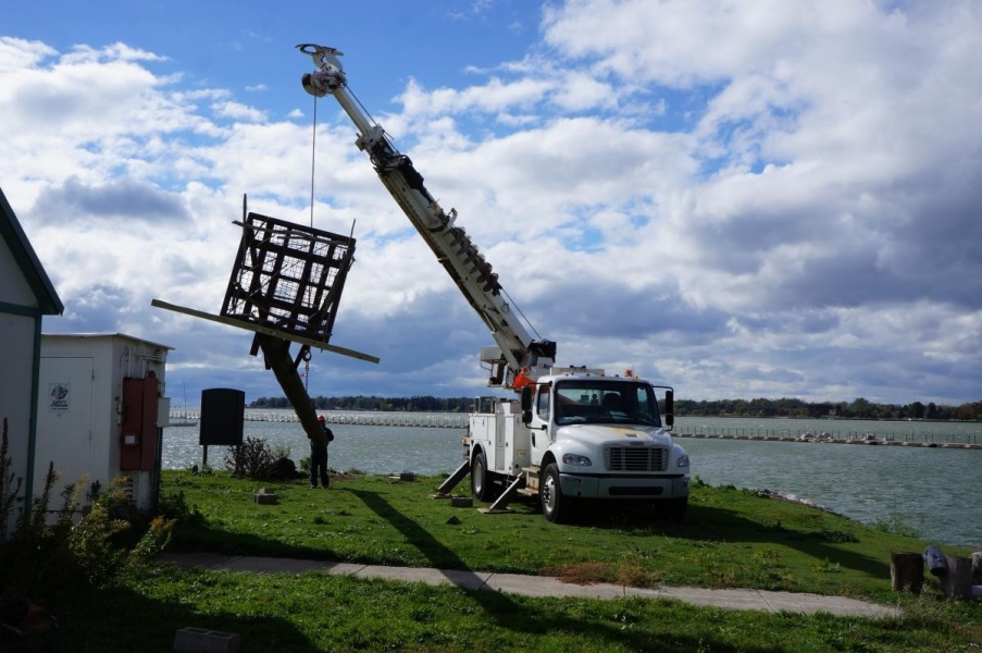 Workers use a truck with a crane to move the osprey pole near the waterfront. The crane is lifting the pole higher while a worker tilts the bottom of the pole toward the ground.