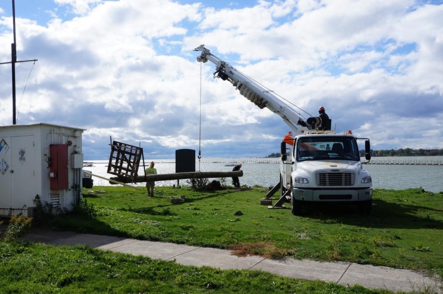 Workers use a truck with a crane to move the osprey pole near the waterfront. The pole is perpendicular to the ground and only a few feet above it, and two workers are guiding it along.