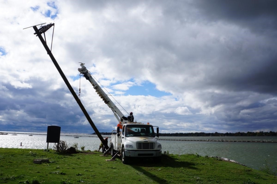 A truck with a crane holds up the osprey pole while two workers put its base in a hole.