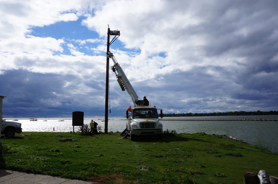 A truck with a crane holds the osprey pole upright in a hole by the waterfront.