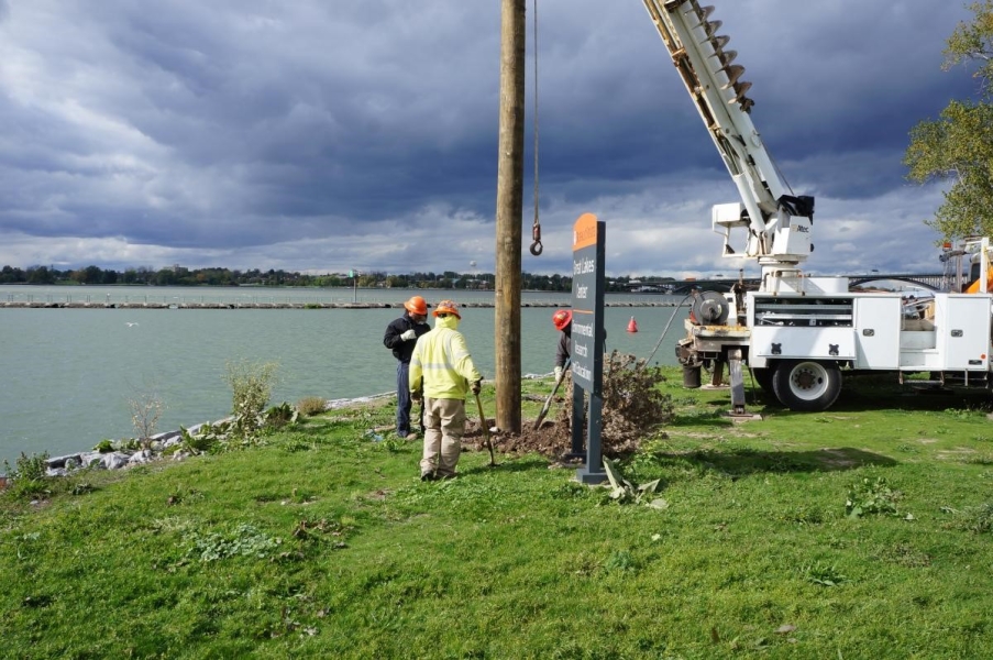 Three workers stand by the bottom of a pole with shovels. They are near the waterfront and there is a sign and a truck behind them.