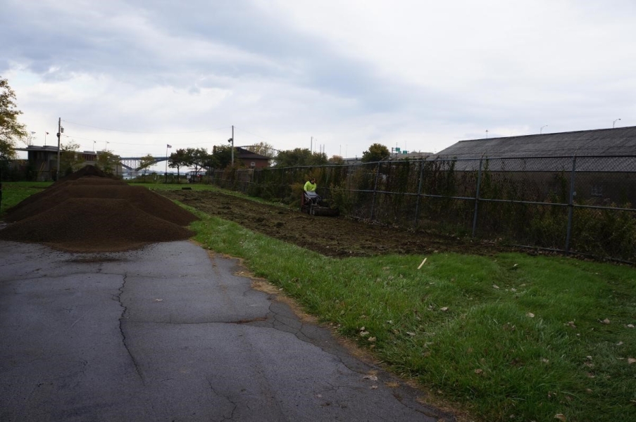 A person pushes a machine in a rectangular area along a fence to remove the grass. There are piles of dirt on a roadway nearby.