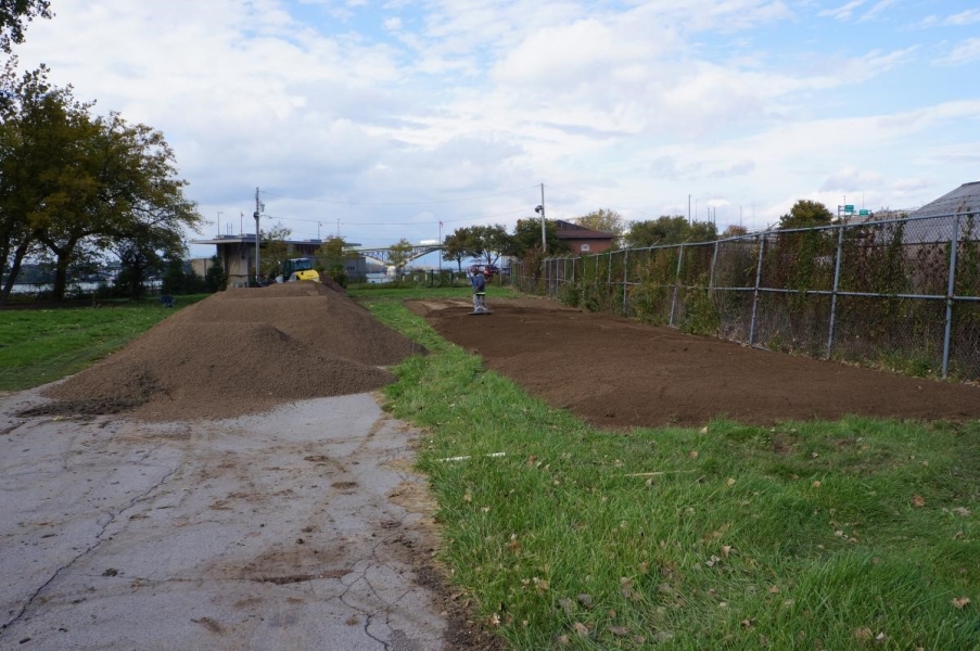 A worker spreads top soil with a rake on a large rectangular plant bed near a fence. There are piles of top soil on the roadway nearby.