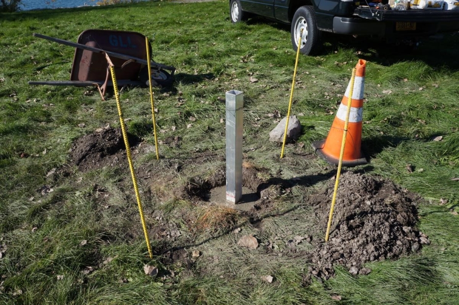 Part of a metal pole in fresh cement in a hole in the ground, with stakes, a traffic cone, and a wheelbarrow turned on its side.