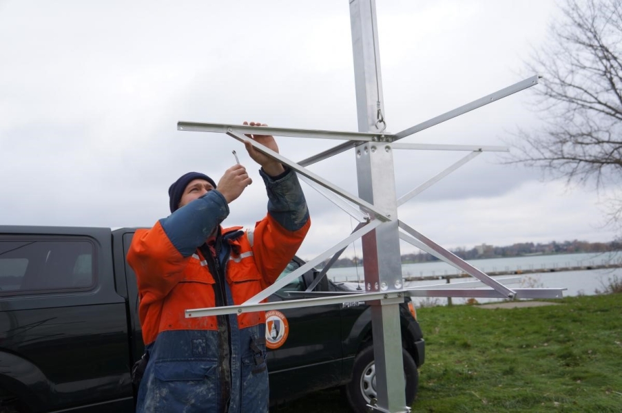 A person in a work suit and knit cap reaches over his head with a small wrench to assemble a metal frame on a pole. There is a vehicle behind him.