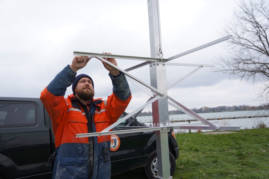 A person in a work suit and knit cap reaches over his head with both hands with a small wrench to assemble a metal frame on a pole. There is a vehicle behind him.