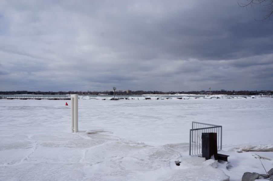 Ice along the shoreline with a metal pipe sticking up through it. There is ice over the breakwall in the distance.