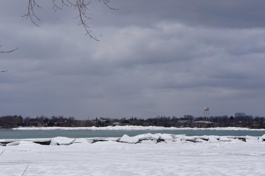 An ice-covered body of water next to an open area of water. There are large pieces of broken ice atop a breakwall between the two bodies of water. On the far shore, there is a giant pile of ice.