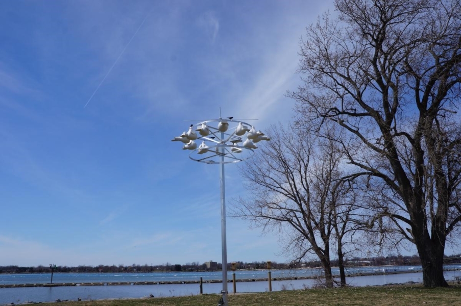 A metal pole with a ring of plastic bird houses at the top in a field by the waterfront. There are a few bird decoys on top of the nesting colony.