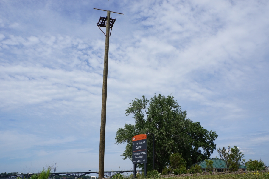 A utility pole with a platform on top stands next to a building sign that says "Great Lakes Center." In the background is a tree, a short building, and a long bridge.