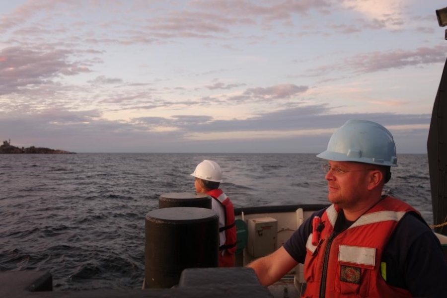 Two people wearing hard hats and life jackets stand at the edge of a large boat.