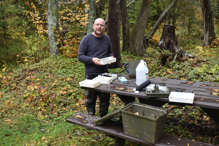 A person stands by a picnic table in the woods in fall. There are plastic pans and jugs on the table, and a plastic pan in his hands.