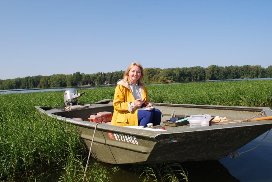 A person sitting in a small boat with sampling equipment, surrounded by short emergent vegetation.