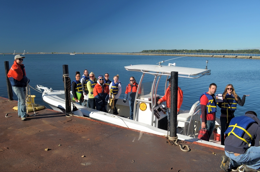 A group of people stand on a fiberglass boat tied up to a dock. Two people on the dock hold the lines.