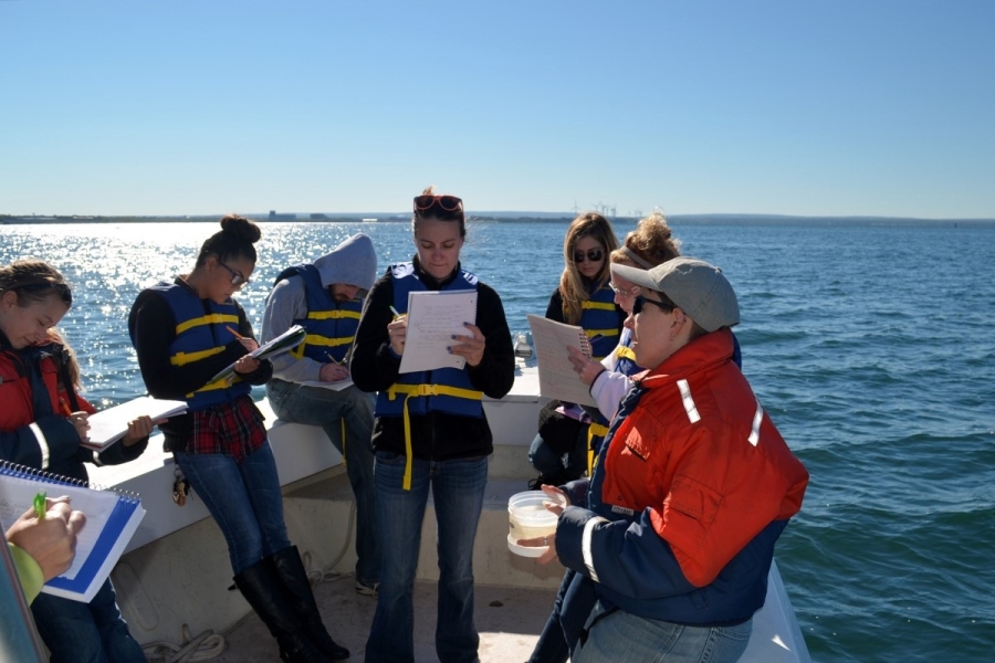 A group of students on a boat take notes while a person holds up a small container of water and gives a lecture