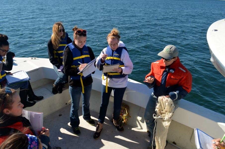 A group of students on a boat take notes while a person holds a conical fine-mesh net and gives a lecture