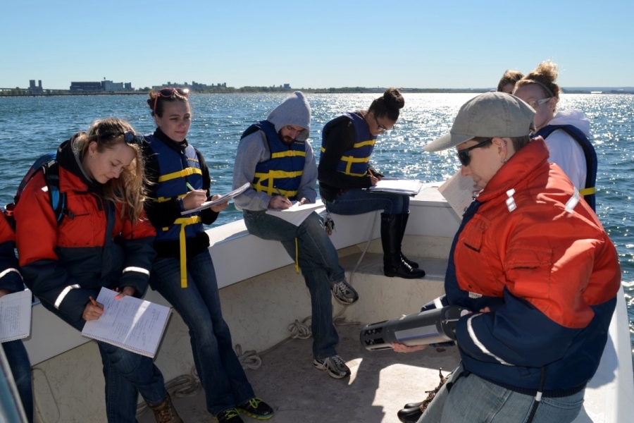 A group of students on a boat take notes while a person holds up a long cylindrical instrument and gives a lecture