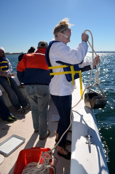 A student standing at the edge of the boat prepares to lower a metal grab sampler over the side