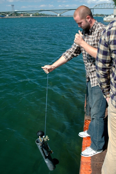 A person lowers a plastic tube on a line into the water from the side of a dock