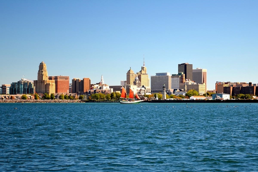 A city skyline with a body of water in front of it on a cloudless day. There is a large sailboat with striking red sails in the foreground.