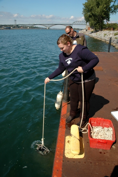 A person lowers a metal grab sampler into the water from the side of a dock