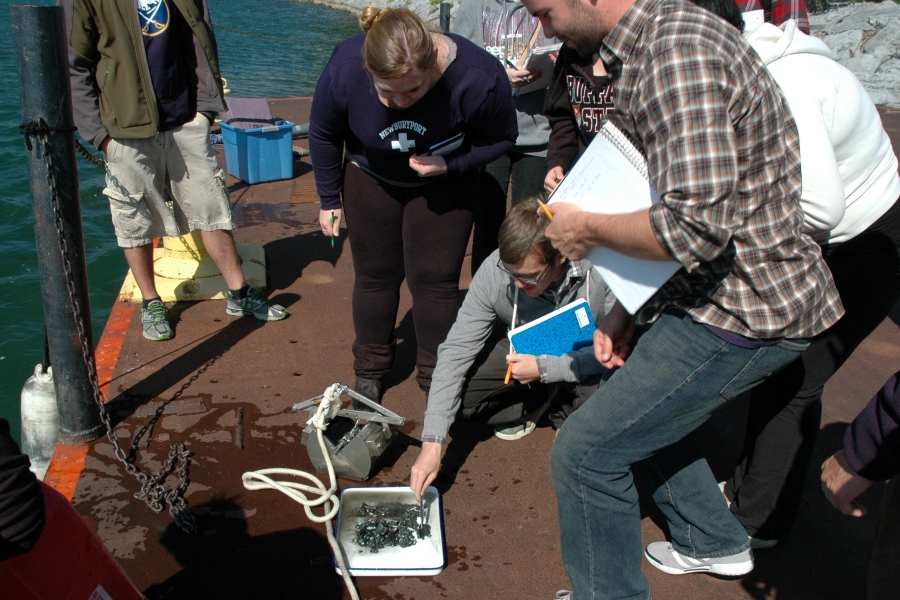 A group of students stand around as someone looks at a muddy sample in a pan. A metal grab sampler sits next to the pan on the dock