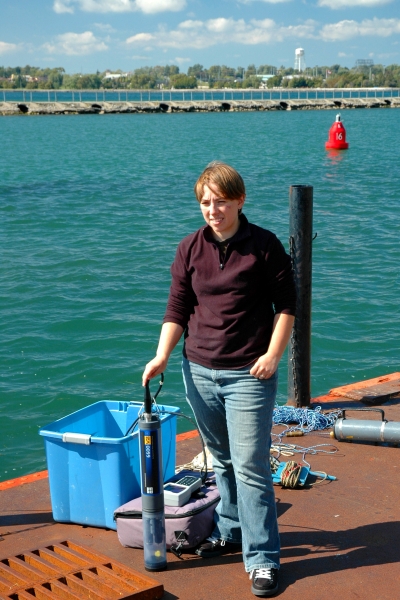 A person stands on a dock by the water with a cylindrical research instrument stood up next to them